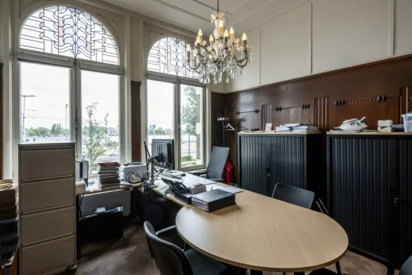 Klassiek ingericht kantoor aan de De Ruijterkade met houten wandpanelen, grote ramen met glas-in-lood en een kroonluchter.